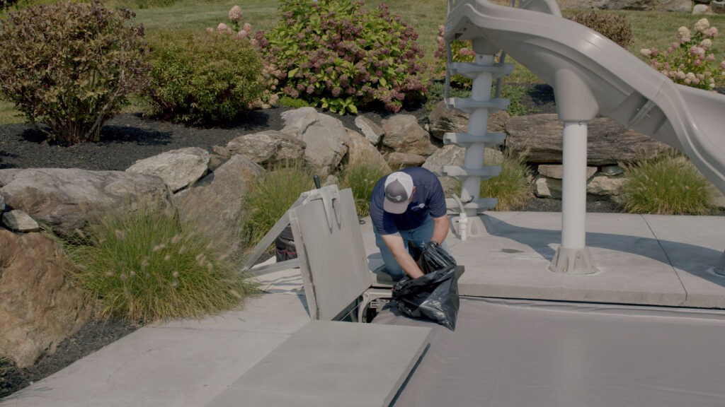 A person wearing a cap is kneeling next to a pool slide, lifting a black trash bag from an open compartment in the poolside concrete area, with rocks and plants in the background.