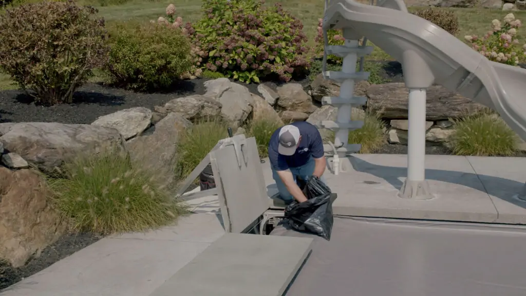 A person wearing a cap is kneeling next to a pool slide, lifting a black trash bag from an open compartment in the poolside concrete area, with rocks and plants in the background.