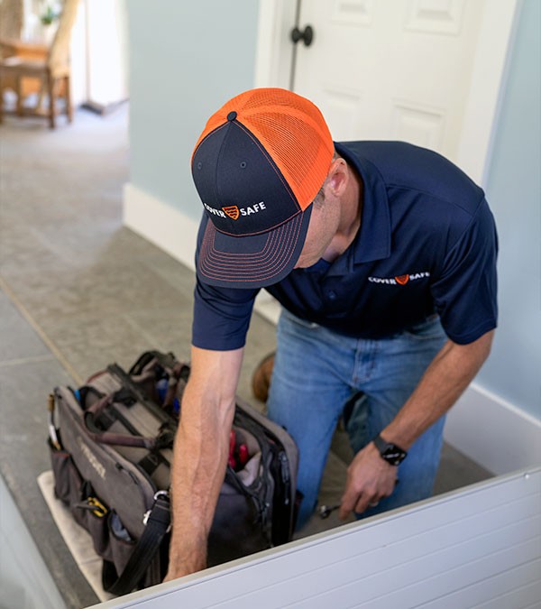 A worker in a navy blue shirt and orange cap kneels indoors, reaching into a tool bag on the floor near a white door. The setting appears to be a residential home.