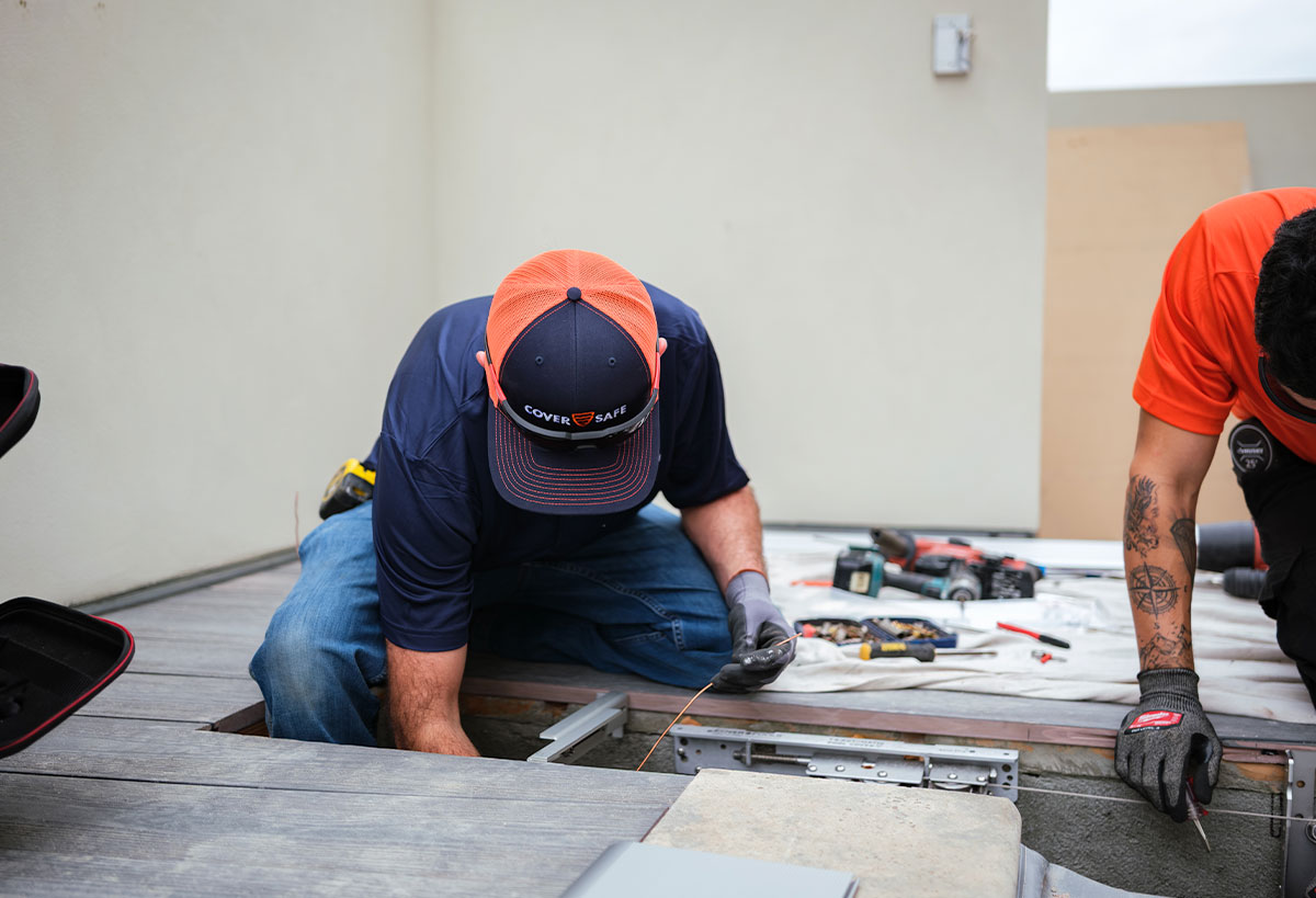 Two workers install decking boards; one kneels while handling wires, and the other works nearby. Tools and supplies are scattered on the floor. Both wear gloves and caps, focused on their task.