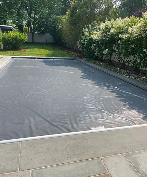 A backyard swimming pool covered with a gray safety cover, surrounded by stone tiles and landscaping with green lawn and bushes in the background.