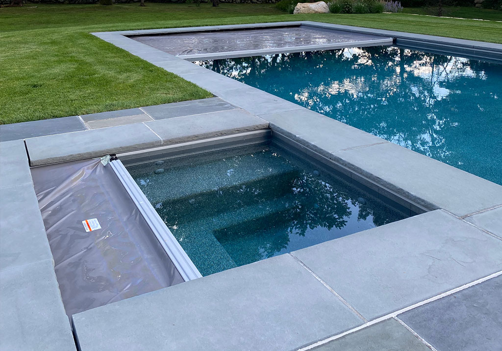 A rectangular hot tub with a partially closed cover is next to a larger swimming pool, both bordered by gray stone tiles and surrounded by green grass.