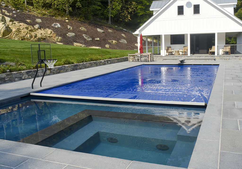 A modern backyard features a rectangular swimming pool with a blue cover, an adjacent hot tub, a small basketball hoop, and a white house with a patio and outdoor seating in the background.