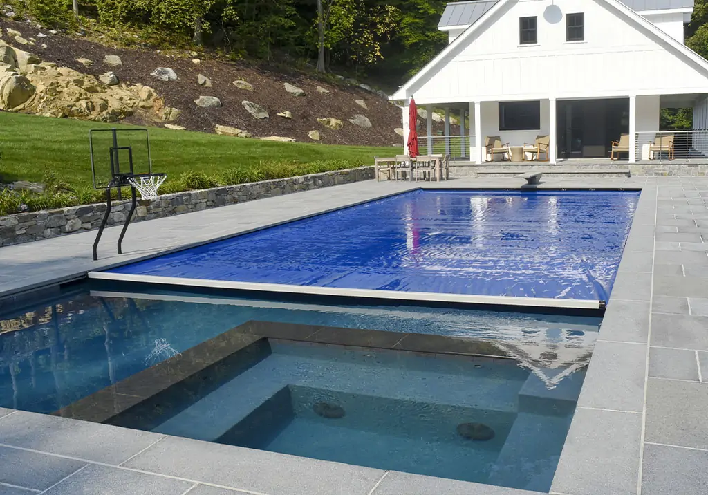A modern backyard features a rectangular swimming pool with a blue cover, an adjacent hot tub, a small basketball hoop, and a white house with a patio and outdoor seating in the background.