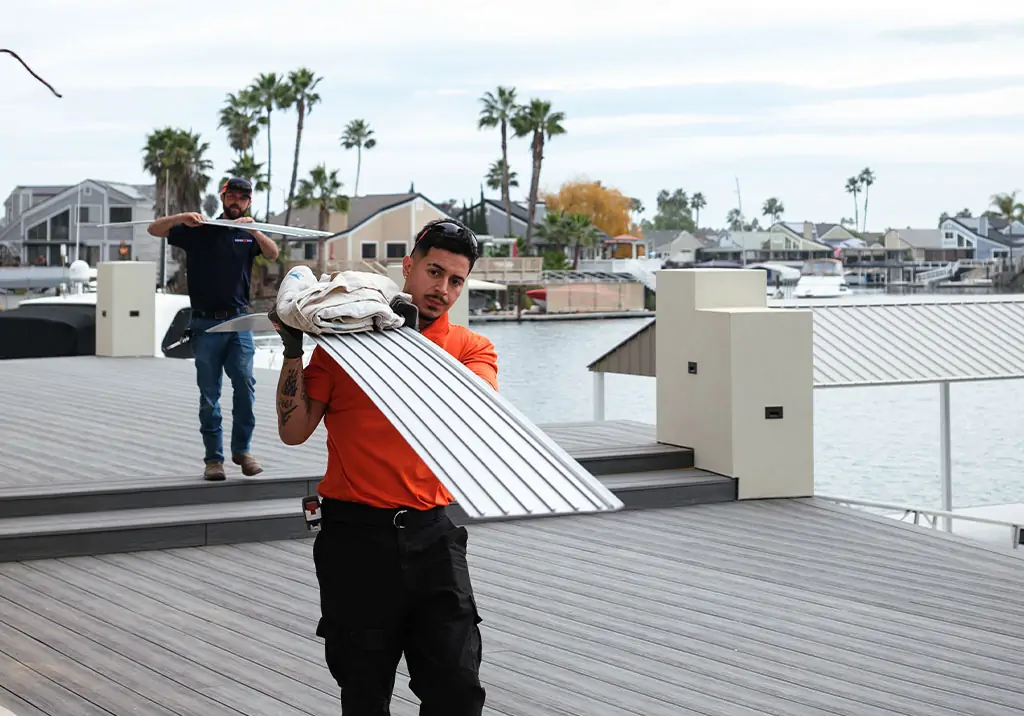 Two men carry long metal planks and supplies on a deck by the water, with houses, palm trees, and docks visible in the background under a partly cloudy sky.