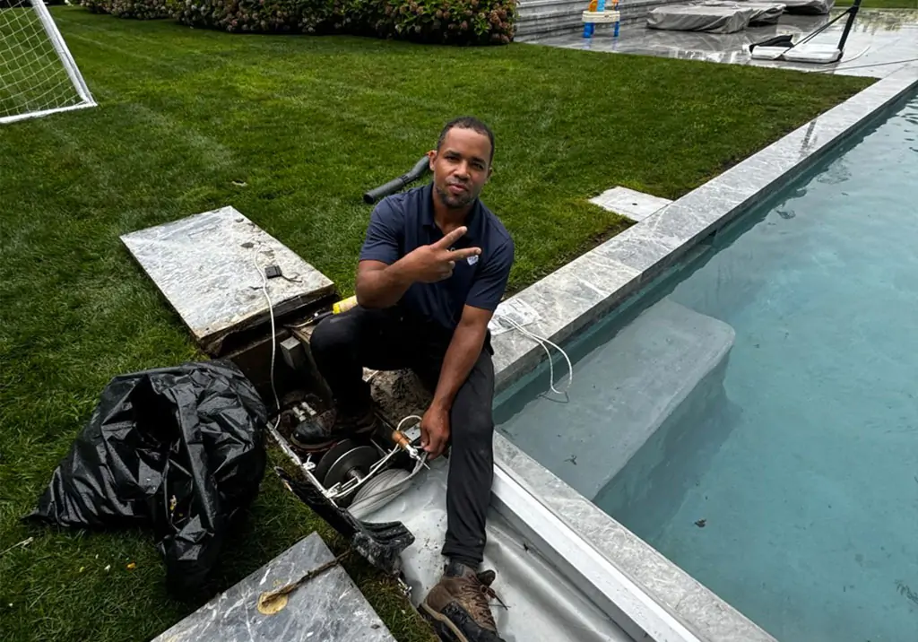 A man sits by the edge of a swimming pool, making a peace sign with his fingers. He is working on pool equipment inside an open compartment. The area is surrounded by grass, with a soccer goal visible in the background.