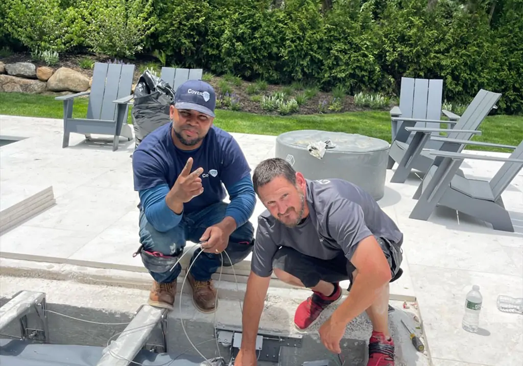 Two men working outdoors on a construction project. One kneels and smiles, making a peace sign, while the other crouches beside equipment. They are surrounded by gray chairs and greenery.