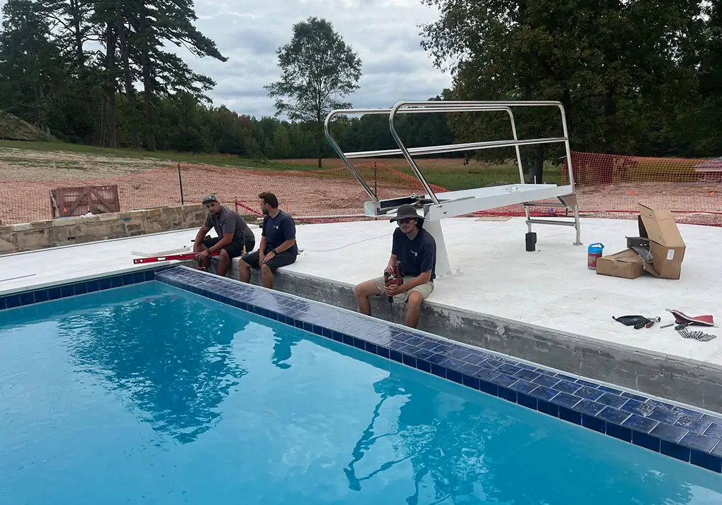 Three men sit on the edge of an outdoor swimming pool near a diving board. They appear to be taking a break from work, with tools and an open box nearby. Trees and a grassy area are visible in the background.