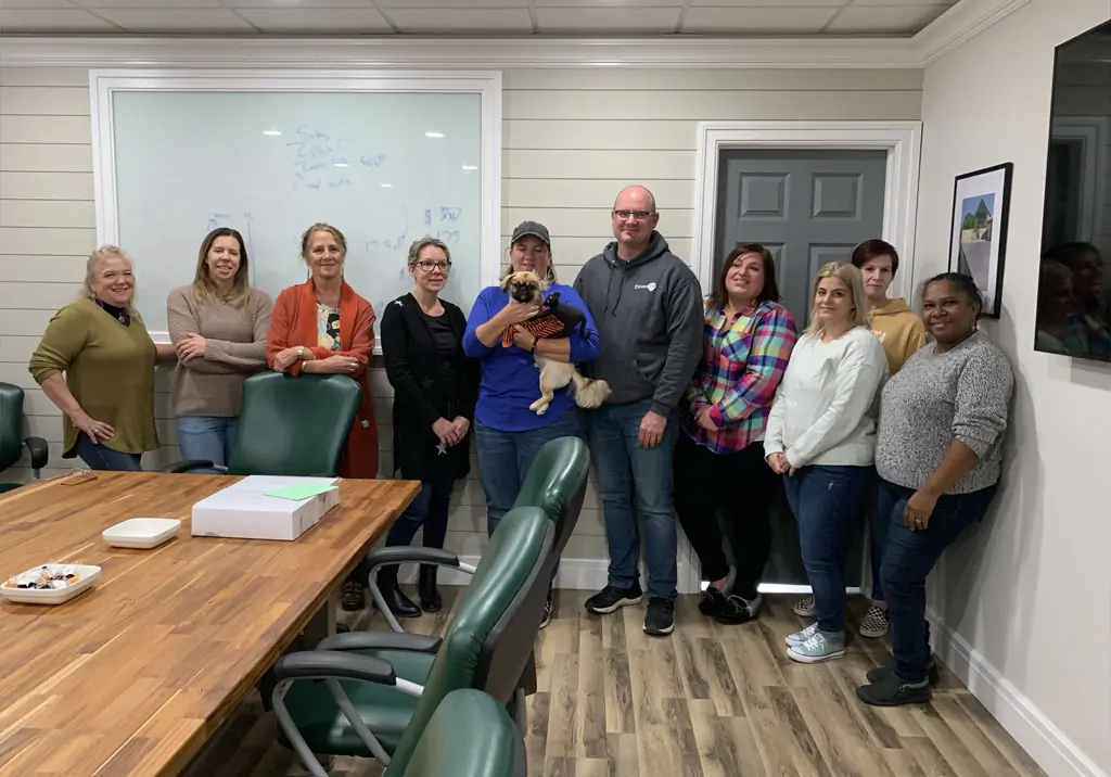 A group of ten people, one holding a small dog, stand and smile together in a conference room with a wooden table, green chairs, and a whiteboard in the background.