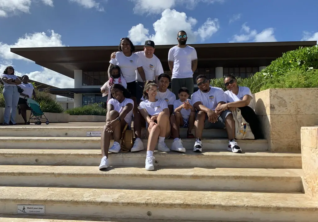 A group of nine people in matching white shirts sit and pose together on outdoor steps, smiling and enjoying a sunny day. A modern building and blue sky with clouds are visible in the background.