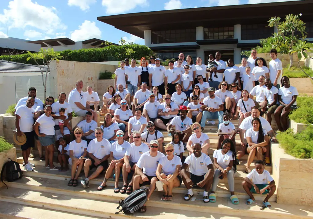 A large group of people wearing matching white shirts pose together on outdoor stone steps, smiling at the camera, with greenery and modern buildings in the background under a blue sky.