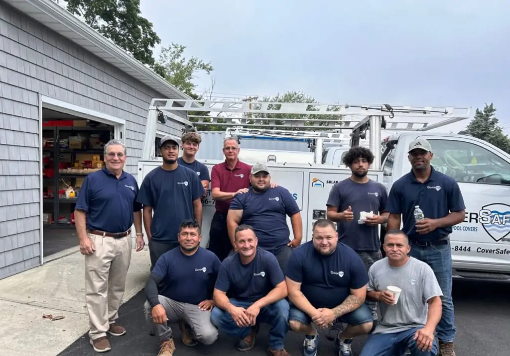 A group of eleven men, some standing and some kneeling, pose for a photo in front of a work truck and a garage. Most are wearing navy blue shirts, and a few hold drinks. The setting appears to be a driveway.