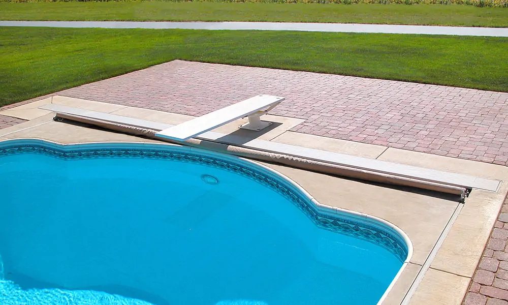 A backyard swimming pool with clear blue water, a curved edge, a white diving board, and a retractable pool cover partially open, bordered by a stone patio and green grass.