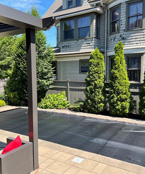 A backyard with a covered swimming pool, patio seating with red cushions, and tall green shrubs along a wooden fence beside a gray house with ornate siding.