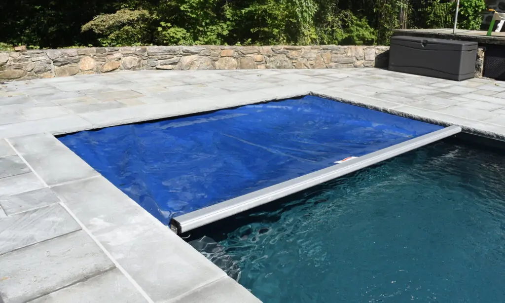 A rectangular pool with a partially closed blue safety cover, surrounded by gray stone tiles and a stone wall, with trees and a storage box in the background.