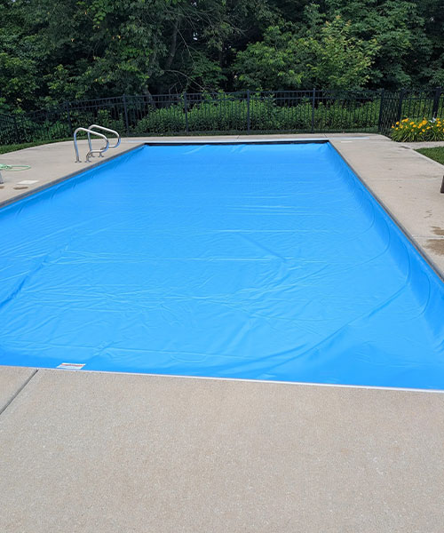 A rectangular outdoor swimming pool covered with a bright blue pool cover, surrounded by a concrete deck and greenery, with a metal pool ladder visible at one end.