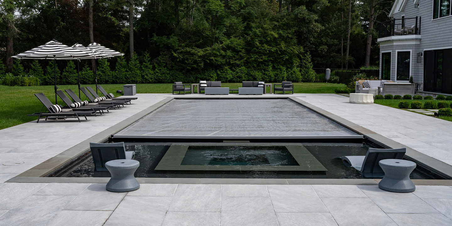 Modern backyard with a covered rectangular swimming pool, built-in hot tub, lounge chairs with striped pillows, patio seating, and black-and-white striped umbrellas, surrounded by greenery and trees.