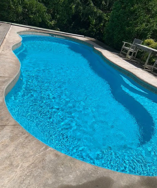 A bright blue, kidney-shaped swimming pool surrounded by a concrete deck, with green trees and patio furniture in the background on a sunny day.