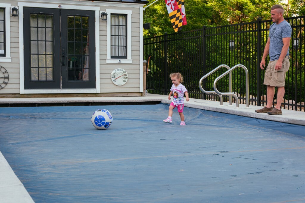 Automatic pool cover closed over a backyard swimming pool, with a child walking on surface of the cover to retrieve a ball